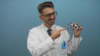 Adam stüdyoda mavi arkaplan ile phoropter işaret ediyor; güven profesyonellik hassasiyet uzmanlığı.