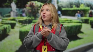 Kırmızı yelek giyen gönüllü bir kadın, canlı bir park ortamında tutkulu bir şekilde jest yapıyor ve açık hava toplum hizmetine olan düşkünlüğünü ve bağlılığını vurguluyor..