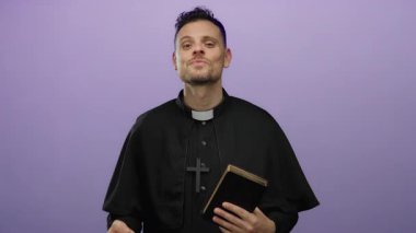 İspanyol rahip İncil 'i işaret ediyor, siyah papaz kıyafeti giyiyor, izole edilmiş mor bir arka plana güvenerek gülümsüyor..