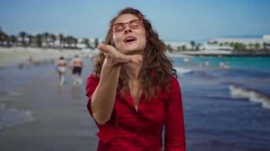 Plajda kırmızı tişörtlü ve gözlüklü bir kadın açık gökyüzünün altında deniz kenarında bir arka planda kameraya öpücük yolluyor..