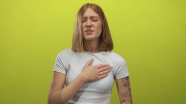 Sarı stüdyo duvarına karşı göğsünü tutan beyaz tişörtlü genç sarışın kadın kederli bir ifadeyle; kalp krizi ağrısı endişesi.