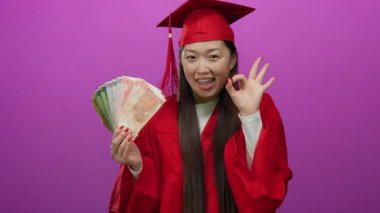 Kırmızı mezuniyet elbisesi içindeki kadın, parlak pembe bir arka plana karşı neşeli bir ifadeyle Filipin pesoları tutuyor, başarıyı ve başarıyı vurguluyor..
