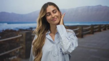 Genç sarışın kadın boynuna dokunur ve deniz kıyısındaki bir kaldırımda ufka bakar..