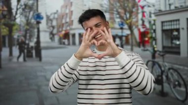 Çizgili süveter içinde gülümseyen genç adam, arka planda canlı şehir hayatı ve eğlencesini yakalayan bisikletli ve hareketli şehir caddesinde kalp hareketi yapıyor..