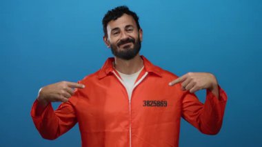 Turuncu hapishane tulumlu İspanyol sakallı adam mavi arka planda güvenle poz veriyor..