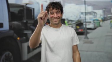 Şantiyede gülümseyen ve el kol hareketi yapan İspanyol bir adam. Kamyon ve şehir arkaplanı, kişilik ve endüstrinin bir karışımını gösteriyor..