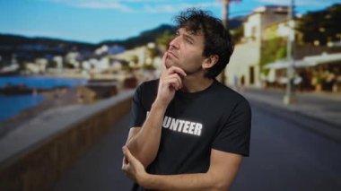 İspanyol bir adam kumsalda denizi tefekkür ediyor. Gönüllü tişörtü giyiyor. Arka planda bulanık bir sahil kasabasıyla dalgın dalgın poz veriyor..