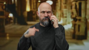 Müzede sakallı, telefonda konuşan kel bir adam evde el hareketleriyle hayal kırıklığını gösteriyor. Stres iletişimi kurarken kafkasyalı erkek dövmesi yaptırıyor..