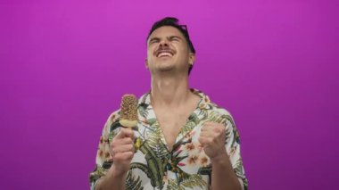 İspanyol asıllı genç adam pembe arka planda, çiçekli gömlekli ve modern tarzda neşeli yaz kaçışını yansıtan pembe elbisesiyle dondurma keyfine varıyor.