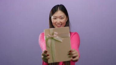 Pembe kazak giyen kadın mor arka planda kurdeleli bir hediye tutuyor. Ayrı bir ortamda neşeli bir ifade ve beklenti sergiliyor..