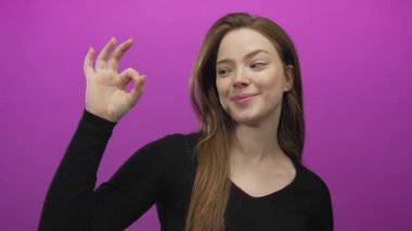 Beyaz bir kadın, iki elini de stüdyoda mor duvara yaslayıp gülümseme ve onaylama işareti gösteriyor..
