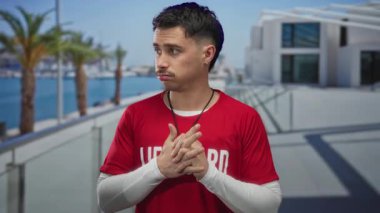 Palmiye ağaçları ve modern mimariyle deniz kenarında gezinen kırmızı tişörtlü genç İspanyol cankurtaran güneşli açık havada odaklanıyor ve hazır bekliyor..