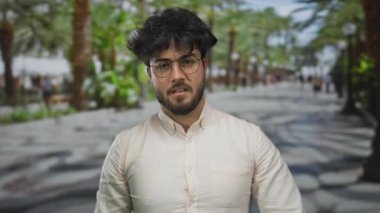 Sakallı ve gözlüklü İspanyol genç adam avuç içi çizgili bir sokakta duruyor, açık bir gömlek giyiyor, bulanık yeşil bir şehir ortamının ortasında..