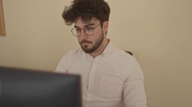 Modern bir ofiste özenle çalışan sakallı ve gözlüklü genç İspanyol bir adam bilgisayar ekranına odaklanmış, kapalı alanda profesyonel ve üretken bir atmosfer yaratıyor..