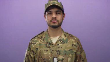 Kamuflaj üniformalı İspanyol asker künyesiyle mor stüdyoda kamerayı işaret ediyor..