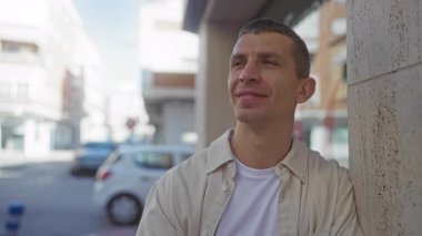 Güneşli bir günde şehir sokaklarında duvarın karşısında gülümseyen bir adam, şehir ortamında iyimserlik ve memnuniyet duygusunu resmediyor..