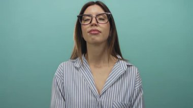 Turkuaz stüdyoda çizgili gömlek ve gözlük takan kadın hafif bir gülümsemeyle gözlerini kapatıyor..