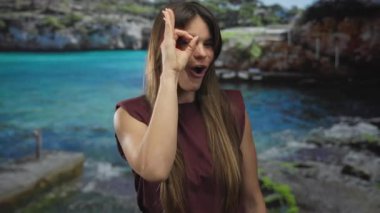 Genç İspanyol kadın, güneşli bir sahilde arka planda turkuaz denizle dikilirken gözünün önünde hoş bir jest yapıyor neşeli yaz titreşimleri sergiliyor..