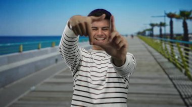 Genç adam elleri ile bir çerçeve hareketi yapıyor, açık bir gökyüzünün altında mavi bir deniz ve palmiye ağaçlarının yer aldığı bir gezinti güvertesinde gülümsüyor..