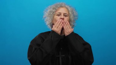 Gri saçlı, rahip kıyafeti giyen bir kadın, sıcak ve nazik bir ifadeyle mavi arka plana öpücük yolluyor..