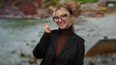 Genç bir kadın deniz kenarında gülümserken el hareketi yapıyor bulanık plaj ve okyanus geçmişiyle..