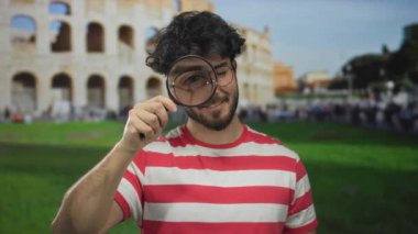 Güneşli bir günde Roma Kolezyumu 'nun önünde elinde büyüteç tutan sakallı genç bir adam.