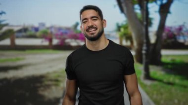 Genç adam gülümsüyor ve gözlerini bir açık hava parkında örtüyor, ağaçların ve çiçeklerin bulanık arka planına karşı mutluluk ve neşe taşıyor.