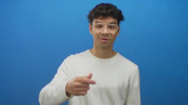 Beyaz uzun kollu tişörtlü genç İspanyol adam mavi stüdyoda el kol hareketi yapıyor; dostça selamlaşma iletişimi.