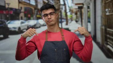 Önlüklü ve gözlüklü genç adam şehir caddesini işaret ediyor. Arka planı bulanık, canlı şehir manzarası var..