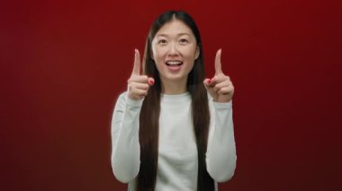 Kadın, canlı kırmızı bir arka planı işaret ederek gülümseyen, uzun saçlı ve beyaz gömlekli olumlu bir jest yapıyor..