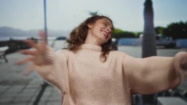 Güneşli bir şehir caddesinde neşeyle gülümseyen bir kadın kollarını açmış hayatı kucaklıyor ve açık havada rahat bir süveter giyiyor neşeli bir şehir ortamında..