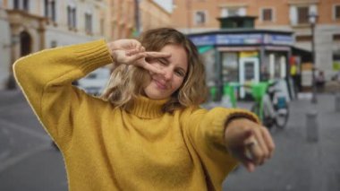 Sokak sahnesinde gülümseyen genç sarışın yolcunun yanında barış jesti yapan ve şehir ortamında bisikleti işaret eden kadın el ele tutuşuyor..
