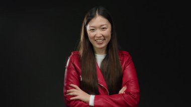 Kırmızı ceketli bir kadın koyu arkaplana karşı güvenle gülümsüyor, kollarını kavuşturarak güzellik ve tarz yayıyor, modern zarafet ve kendine güven sergiliyor..