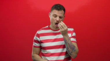 Genç bir adam enerjik bir şekilde kırmızı duvara karşı hapşırıyor, doğal bir tepki gösteriyor, görünür dövme, sıradan çizgili bir gömlek giyiyor, duygularını ve doğallığını ifade ediyor..