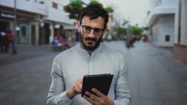 Sakallı İspanyol bir adam işlek bir şehir caddesinde tablet kullanıyor, işine odaklanmış ve meşgul..