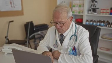 Üniformalı kıdemli erkek doktor hastane kliniğinde bilgisayarla çalışıyor. Arka planda tıbbi aletler ve ekipmanlar var..