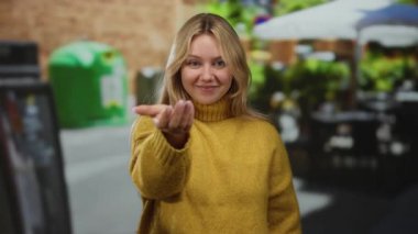 Şehir terasında açık bir kafe ortamında gülümseyen, sarı kazak giyen, sarı saçlı, canlı bir şehir ortamında elini uzatan bir kadın..