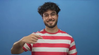 Çizgili gömlek giyen, sakallı ve gözlüklü genç İspanyol bir adam kendini işaret ediyor ve sağlam mavi bir arka plana karşı baş parmak işareti yapıyor..