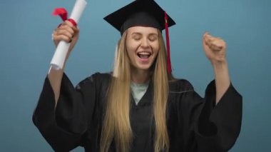 Mezuniyeti kutlayan kadın diplomasını mavi arka planda tutarken neşeli bir ifadeyle akademik önlük ve kep takıyor..