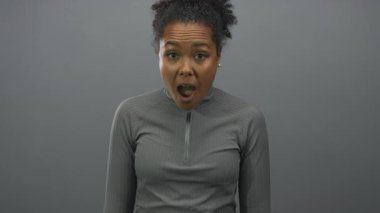 Gri bir stüdyoda, ağzı açık ve kaşlarını kaldırmış, öne doğru eğilmiş genç Afrikalı Amerikalı kadın şaşkınlık....