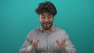 Sakallı İspanyol bir adam mavi arka plan karşısında şaşkınlık ve neşe ifade ediyor, çizgili gömlek giyiyor, duygularını sergiliyor ve modern bir tarzı var..