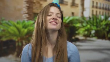 Mavi kazaklı genç beyaz kadın gülümsüyor ve yeşil parktaki parti şapkasını işaret ediyor..