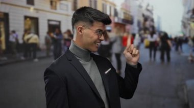 Gözlük takan ve güvenle gülümseyen bir adam arka planda insanların olduğu bir sokak sahnesinde canlı bir şehir atmosferini ön plana çıkarıyor..