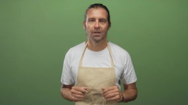 Uzun sarı saçlı, bej önlüklü, beyaz tişörtlü, elleri ve ağzı açık yeşil bir stüdyoda jestler yapan bir adam..