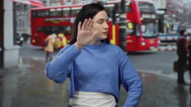 Yağmurlu caddede öfkeli bir şekilde el kol hareketi yapan, arka planda ikonik kırmızı bir otobüsle şehir ortamında hayal kırıklığı ve çatışma gösteren bir kadın.