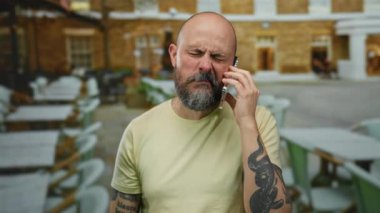 Dışarıdaki kafede sakallı bir adam acilen telefonla konuşuyor. Dövmeli, sıradan bir gömlek giyiyor. Şehir merkezinde arka planda bulanık şehir manzarası ile ilgili endişelerini dile getiriyor..