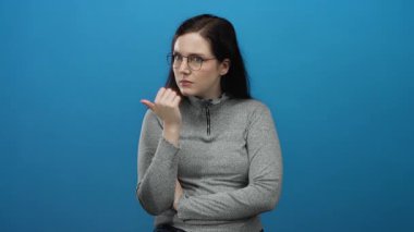 Gözlüklü kadın mavi arka planda gri bir kazak içinde meraklı ve düşünceli bir yüz ifadesini gösteriyor.