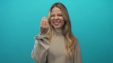 Sarışın kadının eli çimdikli parmaklarıyla turkuaz bir stüdyo arkaplanına jest yapıyor; merak vurgusu.