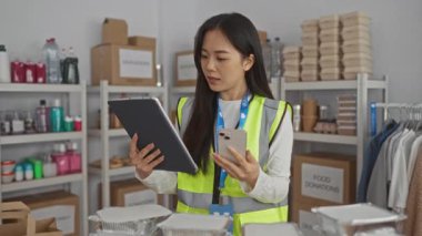 Yansıtıcı üniformalı genç bir kadın yardım merkezinde tablet ve telefon kullanıyor ve raflarda bağış kutuları ve yiyecek malzemeleri var..