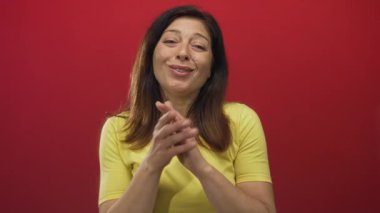 Orta yaşlı, elleri sımsıkı bağlı bir İspanyol kadın ve stüdyoda yumuşak ışıklandırma altında kırmızı duvara karşı nazik bir gülümseme..